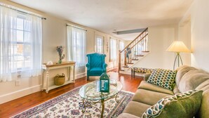 A spacious living room with a blue armchair, a glass coffee table, and a patterned sofa. A staircase leads to an upper level. - Cape Cod Home—Deck, Near Canal & Bike Path (Sandwich)