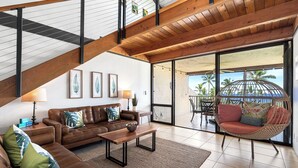 A spacious living area with a brown leather sofa, a wooden coffee table, and a wicker chair. There are framed pictures on the wall, a lamp, and a view of the outdoors through large glass doors.