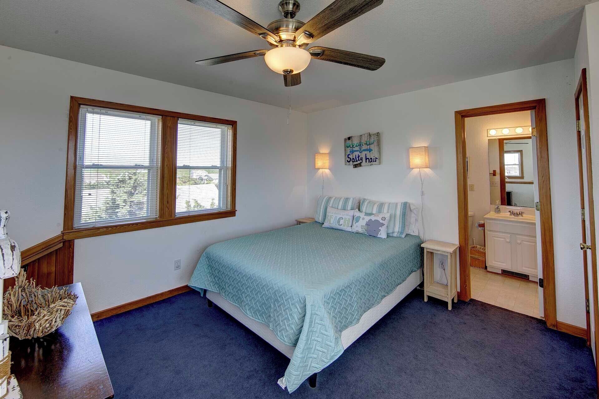 A bedroom with a bed, a ceiling fan, a window, a nightstand, a mirror, and a door leading to a bathroom.