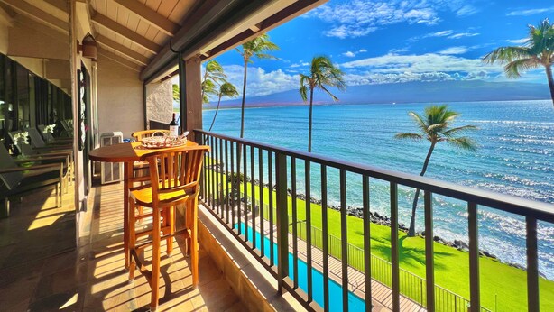 A balcony with a wooden table and chairs, overlooking a pool and a clear view of the ocean with palm trees. - Oceanfront spacious 1bd Maalaea condo (Wailuku)