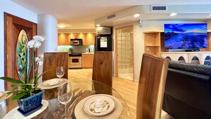 A modern living room with a dining area, featuring a glass table, wooden chairs, a sofa, a TV, and a kitchen in the background. - Oceanfront spacious 1bd Maalaea condo (Wailuku)