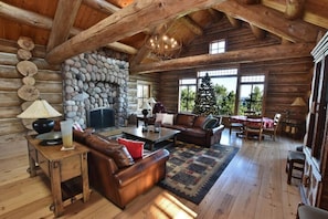 A log cabin interior with a stone fireplace, leather furniture, and large windows offering a view of the outdoors. - Luxury Waterfront Log Home 15 min from Traverse City w/ Dock (Suttons Bay)