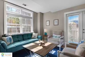 Een woonkamer met een blauwgroene bank, een houten salontafel en een witte fauteuil met een kussen waarop 'BEACH' staat.