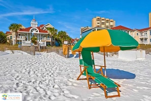 Een strand met een reddershutje en een kleurrijke parasol, een houten hek en gebouwen op de achtergrond.