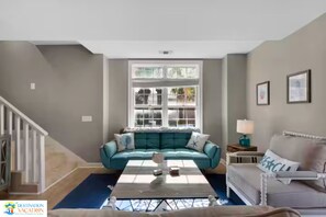 Een woonkamer met een blauwgroene bank, een witte fauteuil, een salontafel en een raam met veel ruitjes.
