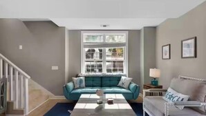 Ein Wohnzimmer mit einem türkisfarbenen Sofa, einem weißen Sessel, einem Couchtisch und einem Fenster mit Blick auf Bäume.