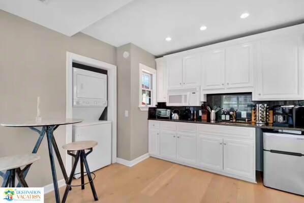 Una cocina moderna con armarios blancos, un refrigerador de acero inoxidable y un comedor con mesa de vidrio y taburetes.