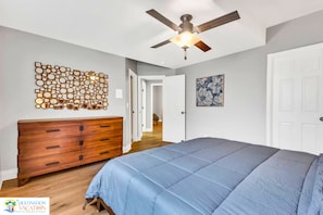Quarto com cama, penteadeira, ventilador de teto e uma obra de arte na parede.