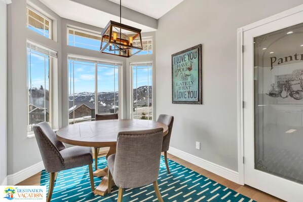 A dining area with a round table, four chairs, a chandelier, large windows, and a view of a snowy landscape.