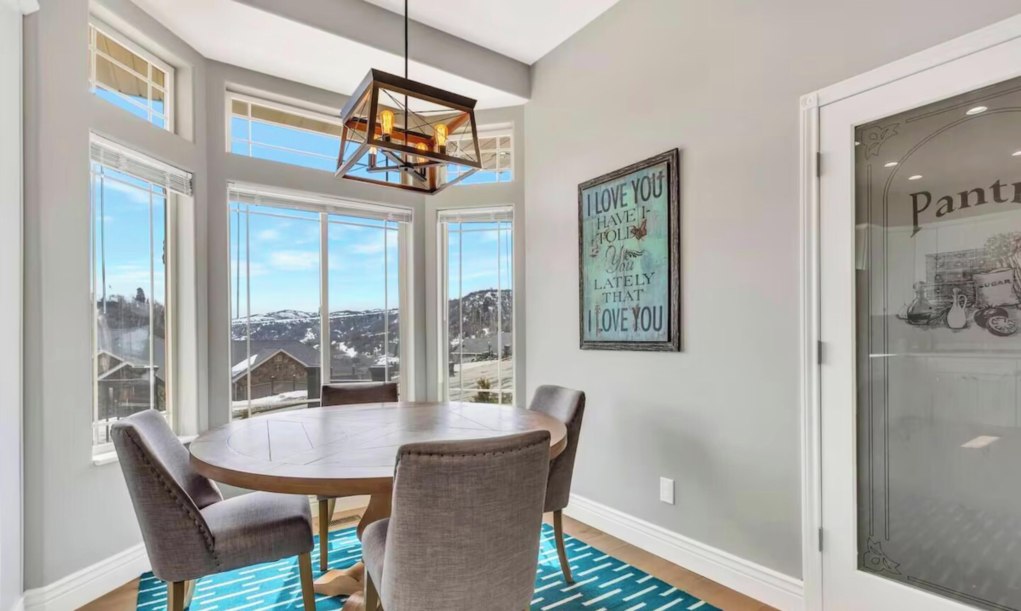 A dining area with a round table, four chairs, a hanging light fixture, and a view of a snowy landscape through large windows.