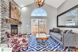 A spacious living room with a stone fireplace, a large sofa, a patterned rug, and a view through a glass door.