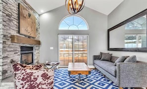 A living room with a stone fireplace, a sofa, a patterned rug, and a large window. - Ultimate Mountain Stay | 6BR Home Near Skiing & Hiking (Mountain green)