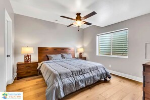 Une chambre à coucher avec un lit, deux tables de chevet avec des lampes, un ventilateur de plafond et une fenêtre avec des stores.