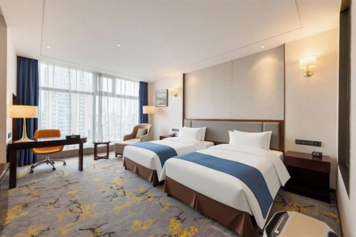 A hotel room with two beds, a desk, a chair, and a view of the city.