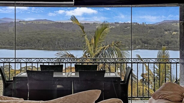 Un balcón con vistas a un lago y colinas, amueblado con una mesa y sillas, rodeado de plantas en macetas.
