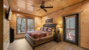 A wooden cabin bedroom with a bed, a ceiling fan, a fireplace, and a window with blinds. - Creekfront Views! Hot Tub/ 2 King Bed En Suites/Secluded/Arcade/Firepit/FastWifi (Pigeon Forge)