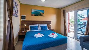 A hotel room with a large bed, a nightstand, a wall-mounted air conditioner, a painting of a coastal scene, and a view of a palm tree through a sliding glass door. - Stone Fence (Sint Willibrordus)
