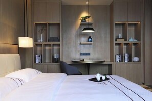 Habitación de hotel con cama, mesita de noche, lámpara, estantes con objetos decorativos y una pequeña mesa con una silla.