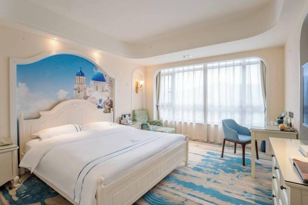 A hotel room with a large bed, a desk, a chair, a TV, and a mural of a blue building.