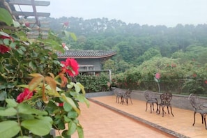 A traditional building with a tiled roof, surrounded by lush greenery and a paved area with chairs.