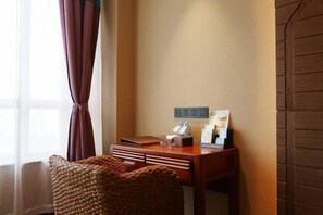 Una habitación de hotel con un escritorio de madera, una silla tejida, una ventana con cortinas y un enchufe en la pared.