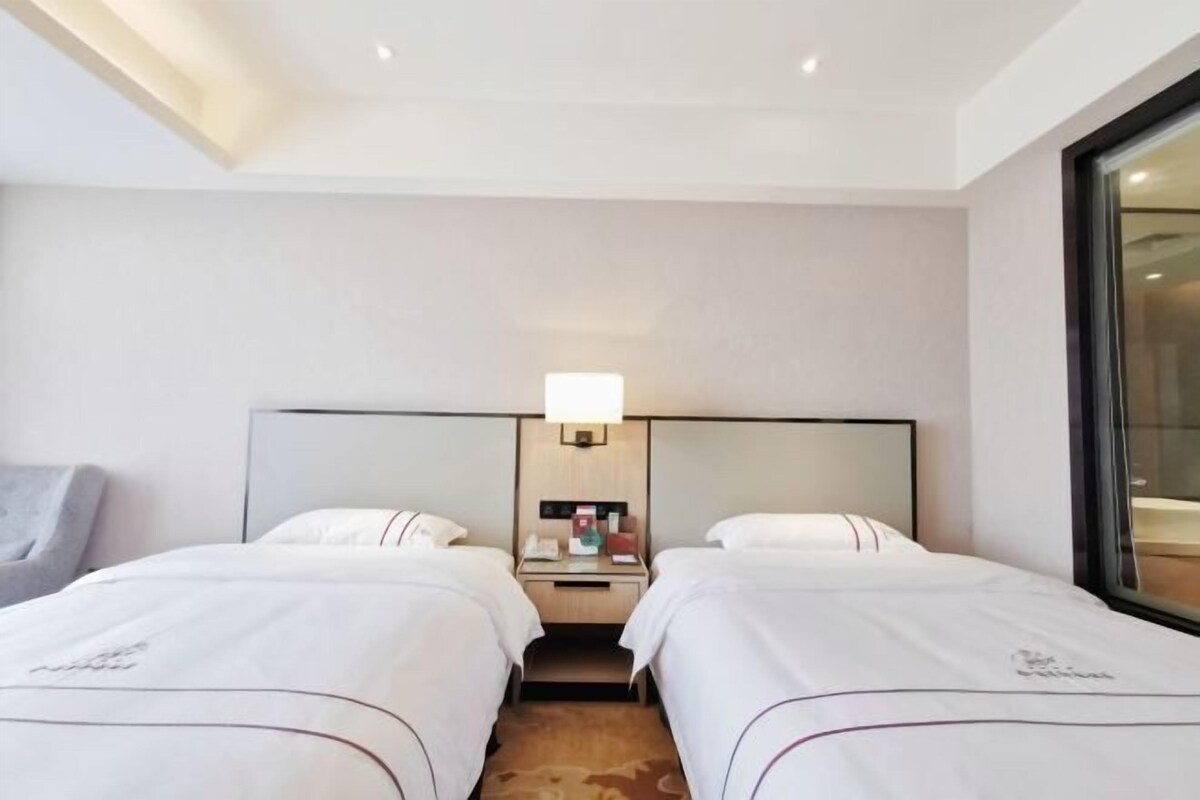 Hotellihuone, jossa on kaksi sänkyä, yöpöytä lampun kanssa ja suuri ikkuna.