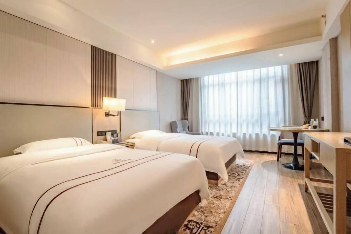 A hotel room with two beds, a desk, a chair, a window with curtains, and a patterned rug.