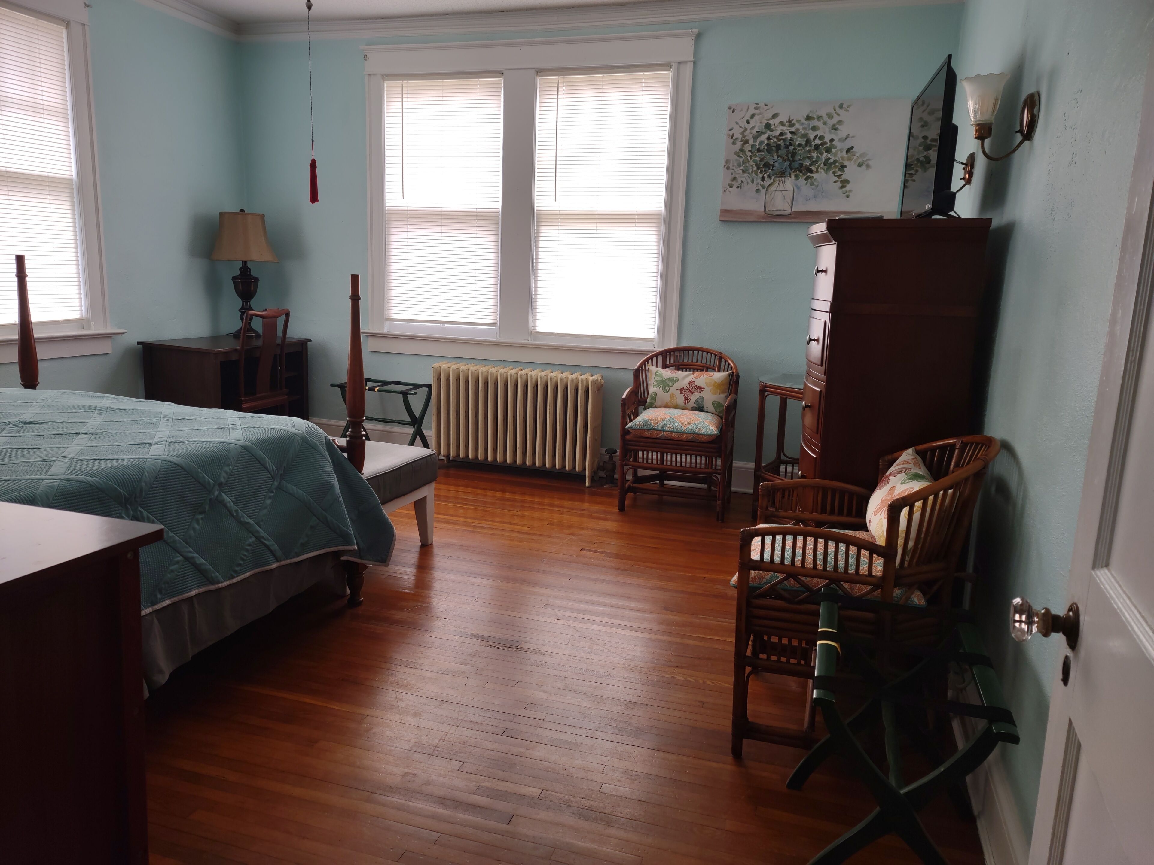 Une chambre à coucher comprenant un lit, deux chaises, une commode, un radiateur et une fenêtre avec des stores.