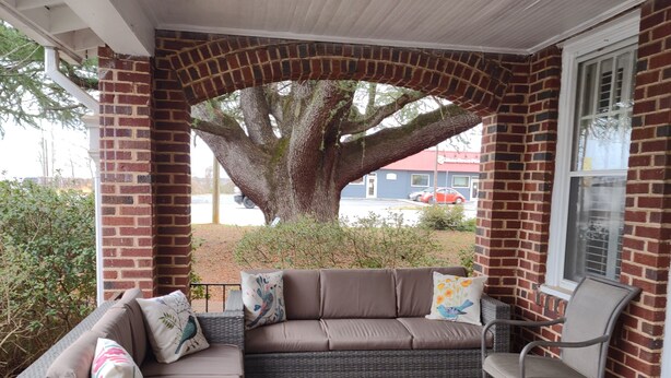 A covered patio with a brick wall, a large tree, a wicker sofa, a metal chair, and a tiled floor. - Welcome To Downtown Pickens close to Clemson and Lake Keowee (Pickens)