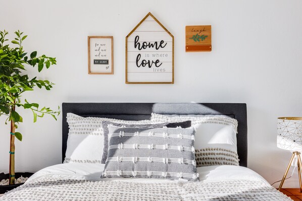 Een keurig opgemaakte slaapkamer met een bed, kussens, een kamerplantje en wanddecoraties, waaronder lijstjes met teksten.