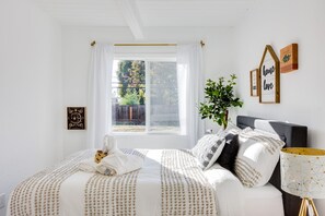 Een slaapkamer met een groot raam, een bed met een wit-beige gestreept dekbedovertrek, een nachtkastje met een lamp, en een muur met ingelijste foto's en een bord.