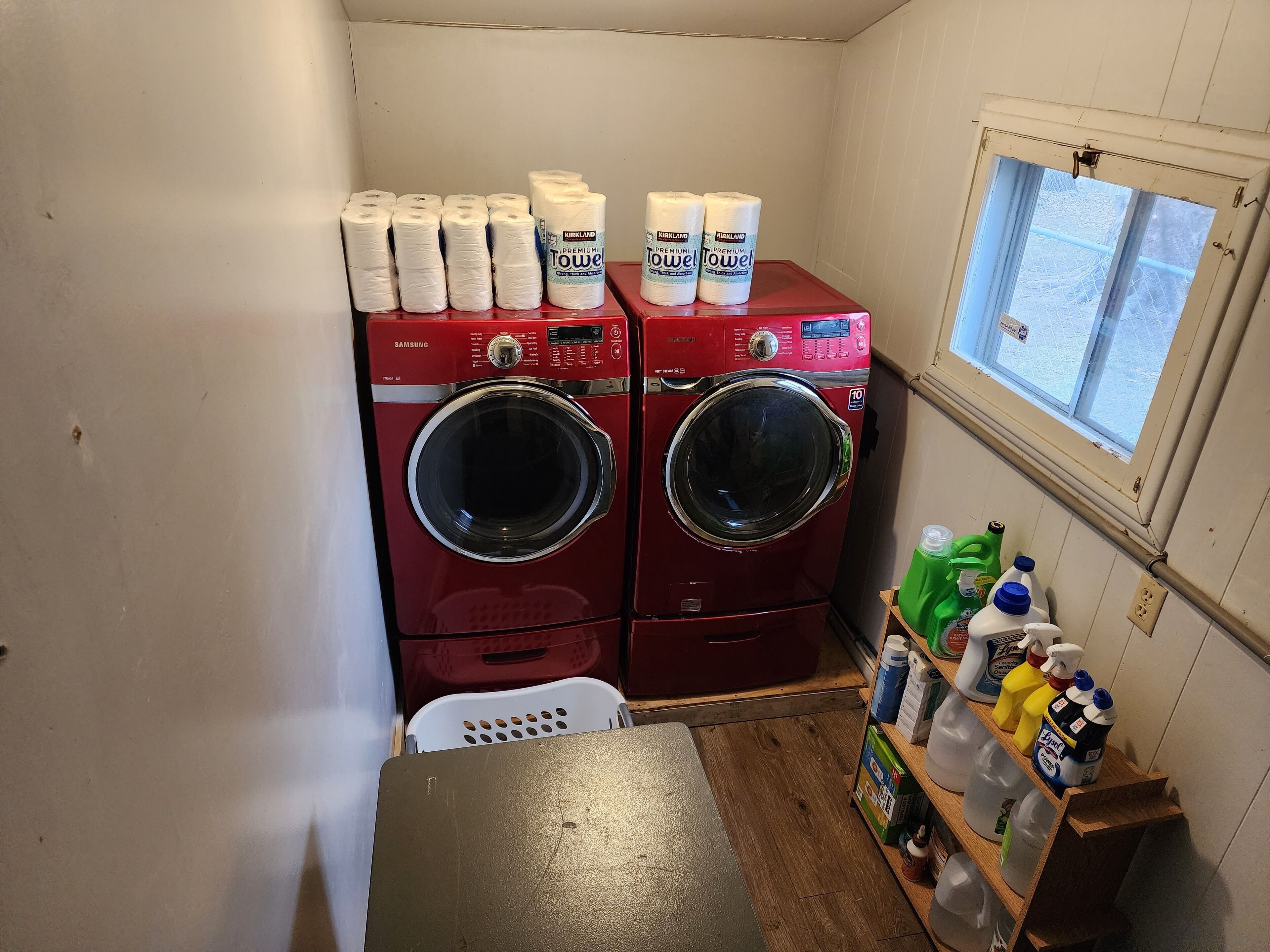 A laundry room with two red Samsung washers and dryers, a window with a view, and various cleaning supplies on shelves.