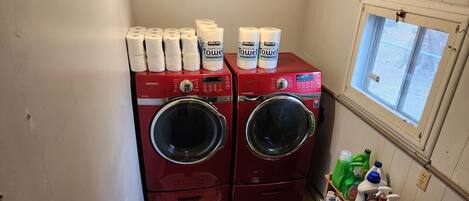 A laundry room with two red Samsung washers and dryers, a window with a view, and various cleaning supplies on shelves.