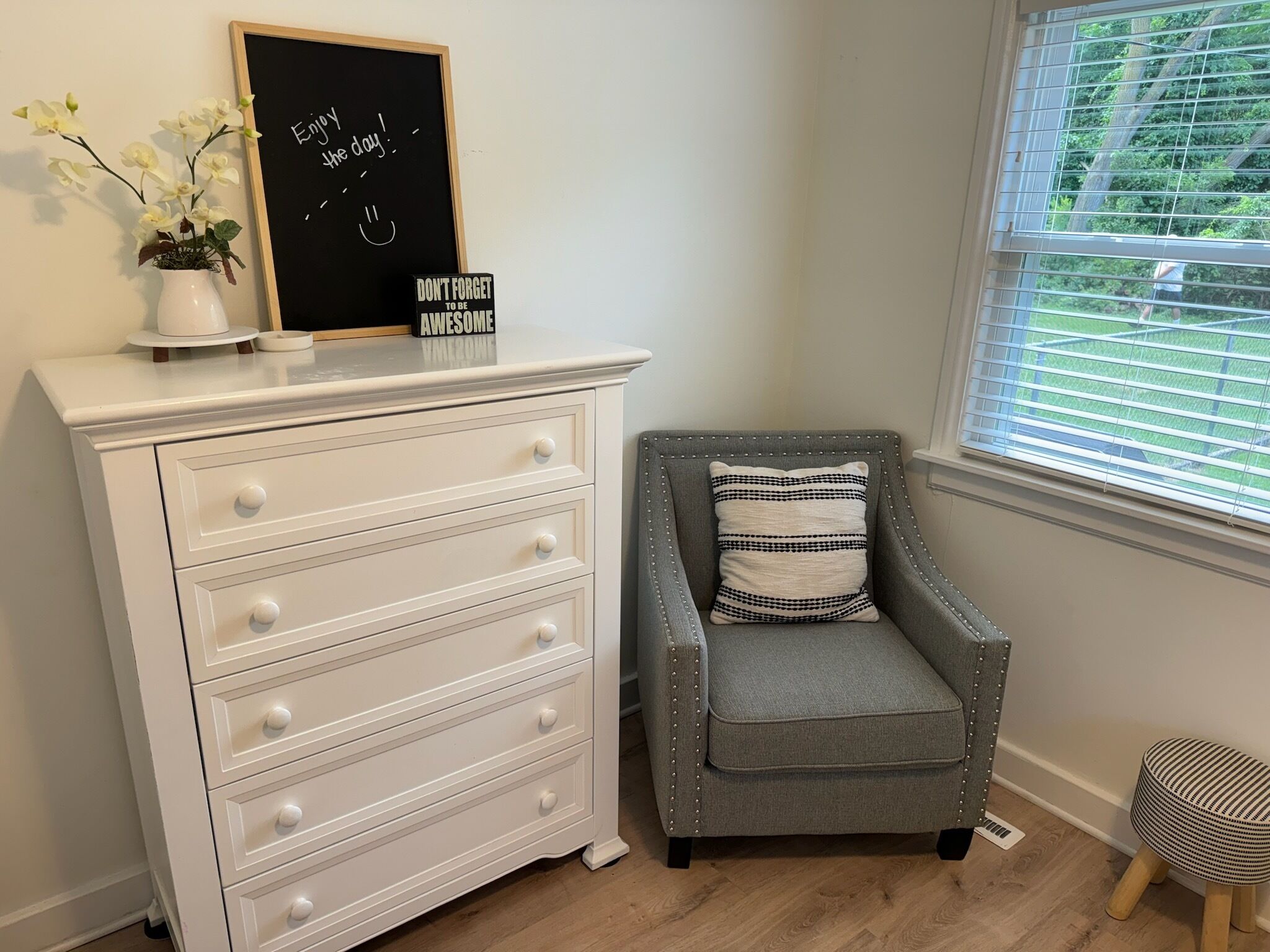 A room with a white dresser, a gray armchair with a striped pillow, a blackboard with motivational writing, and a window with blinds.