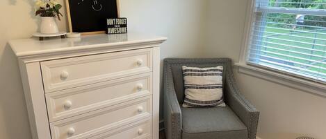 A room with a white dresser, a gray armchair with a striped pillow, a blackboard with motivational writing, and a window with blinds.