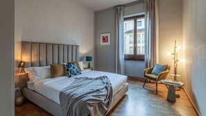 Habitació d'hotel amb un llit gran, una cadira, una taula petita i una finestra amb cortines.