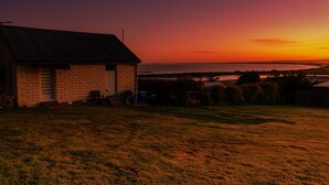 Une maison aux murs en pierre et au toit métallique, située dans une zone herbeuse, offrant une vue sur une étendue d’eau au coucher du soleil.