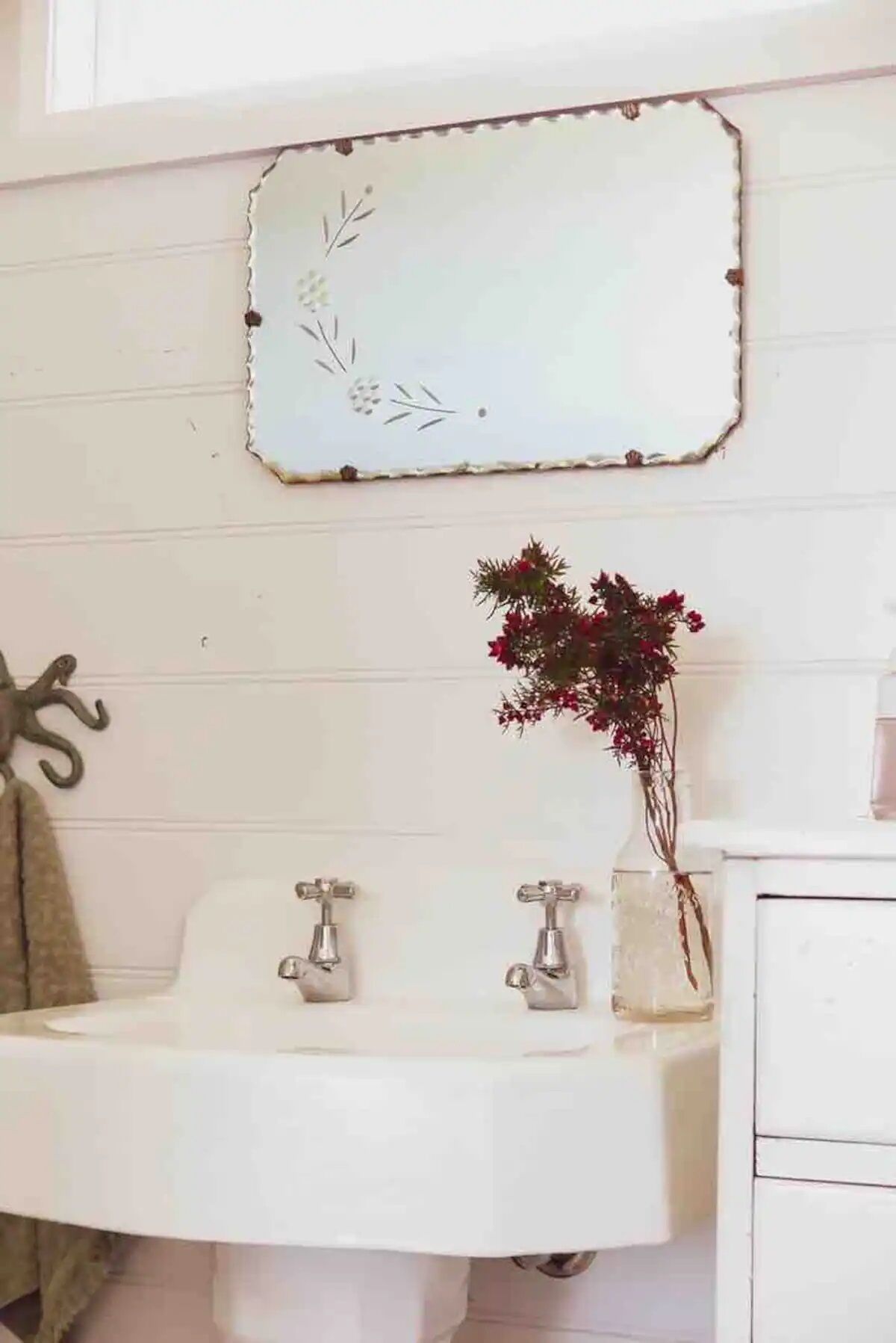 Un baño con un lavamanos blanco, dos grifos, un adorno sobre el lavamanos y un florero con flores rojas.