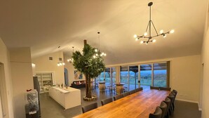 A spacious dining area with a long wooden table and multiple chairs, a central tree, and a view of the outdoors through large windows. - Kesehills Wines Villa (Mount Pleasant)