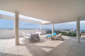 A covered outdoor area with a table and chairs, a sun lounger, and a view of the sea and buildings in the distance. - Holiday apartment mit Klimaanlage und Balkon (Lignano)