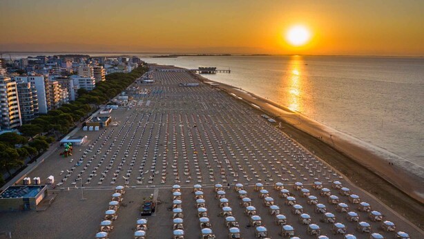 A beach with rows of umbrellas and chairs, a pier in the background, and buildings along the shore.