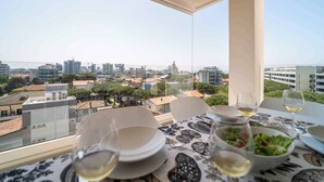 A dining table with plates, glasses, and a salad bowl, set against a cityscape view from a balcony.