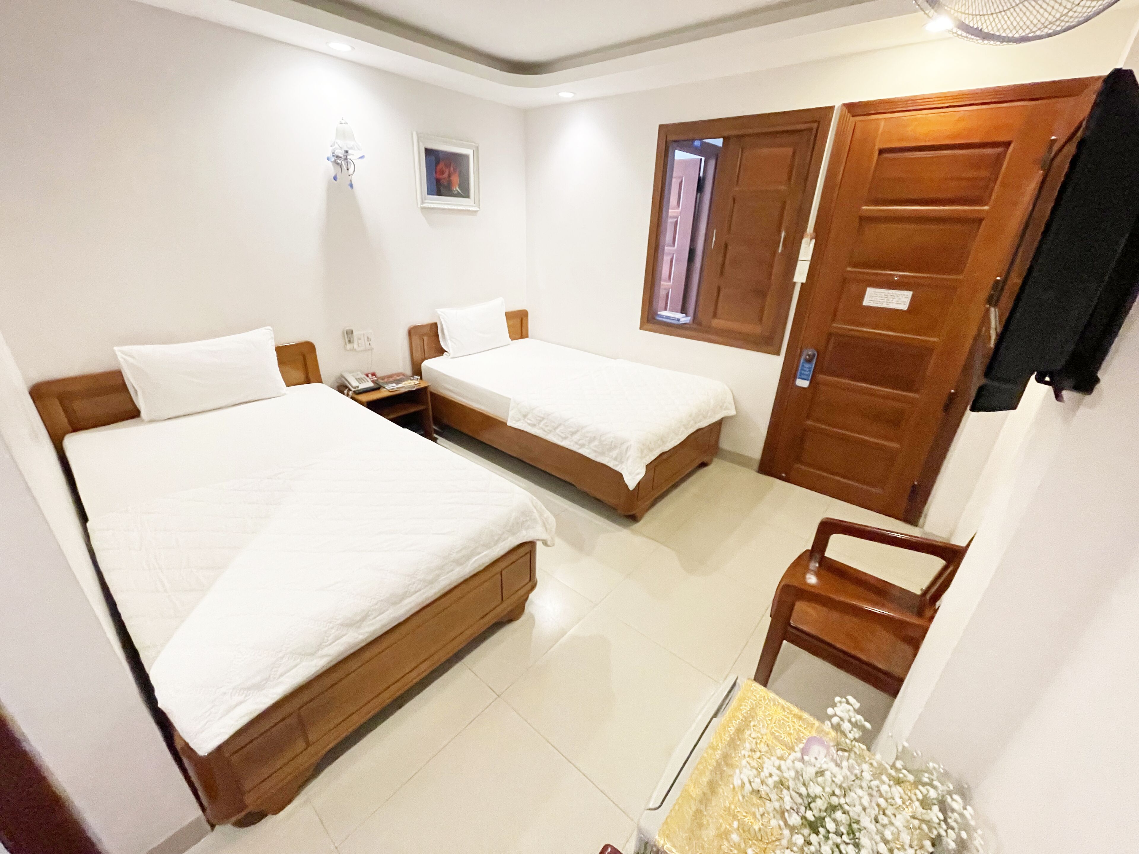 A hotel room with two single beds, a wooden chair, a small table, a wall-mounted fan, a framed picture, and a door with a window.