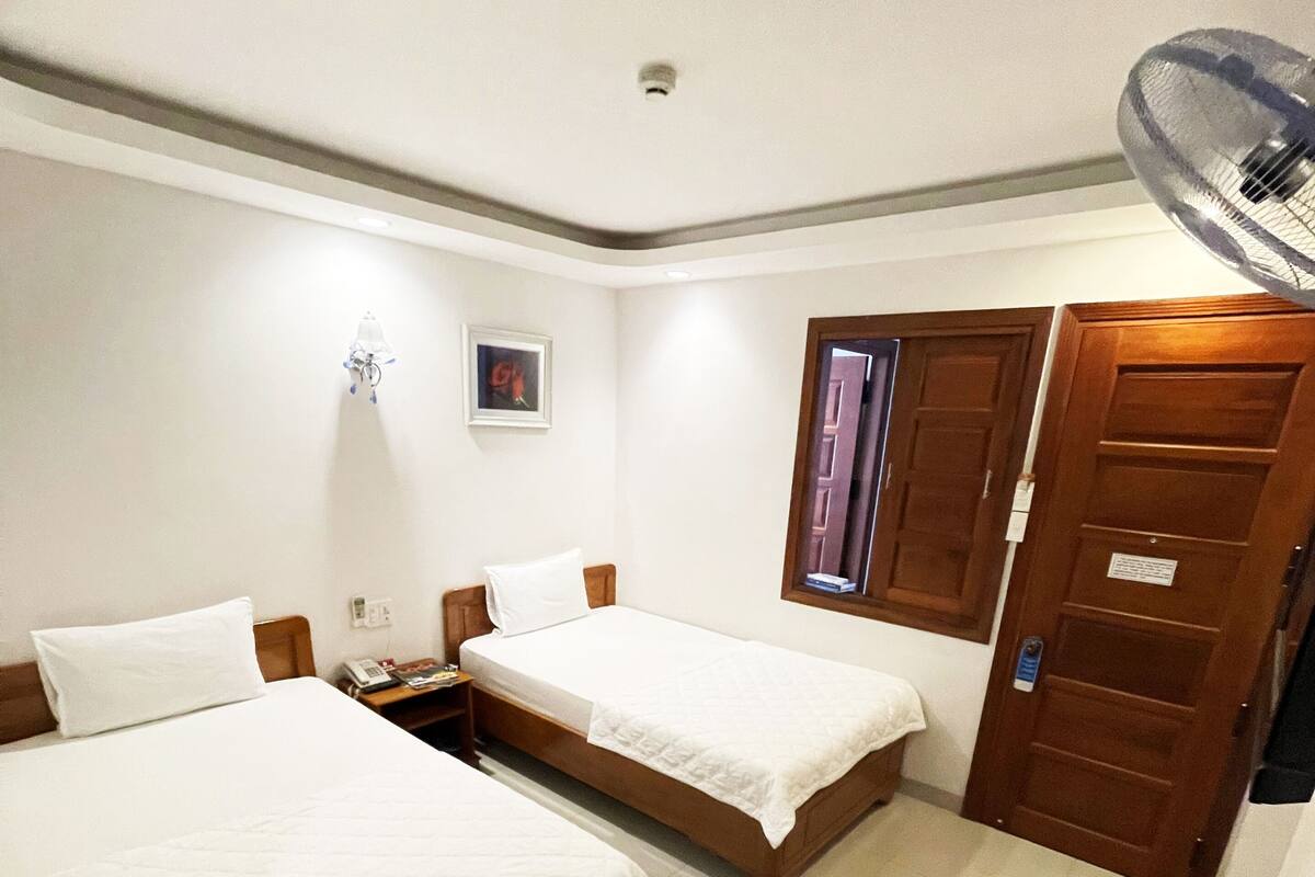 A hotel room with two single beds, a wooden door, a small table with a chair, and a wall-mounted light fixture.