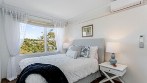 Ein Schlafzimmer mit einem Bett, einem Nachttisch mit Lampe, einem Fenster mit Vorhängen und einer Wandkonsole mit Klimaanlage.
