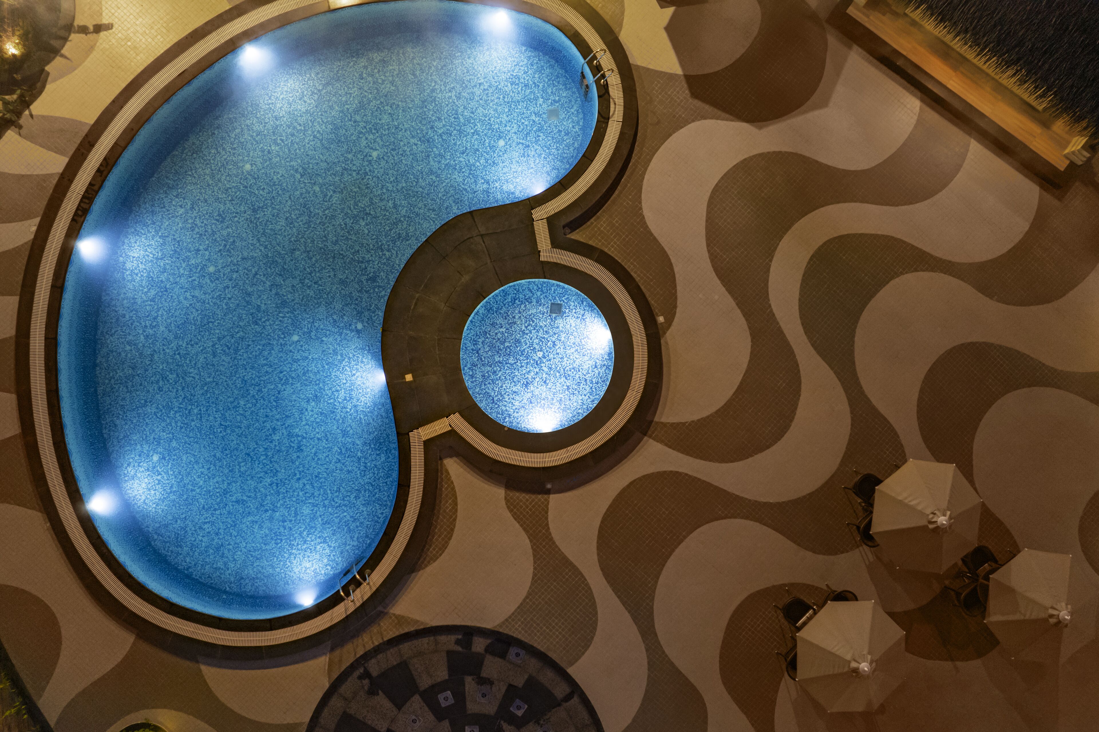 A pool with a unique shape, surrounded by a patterned floor and several umbrellas.