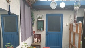 Un petit espace extérieur clos, avec deux portes bleues, un banc en bois, une petite table et une chaise rouge. On y trouve également des plantes et une horloge accrochée au mur.