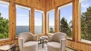 Una habitación acogedora con ventanas amplias, dos sillones, una mesita y vista al agua y los árboles.