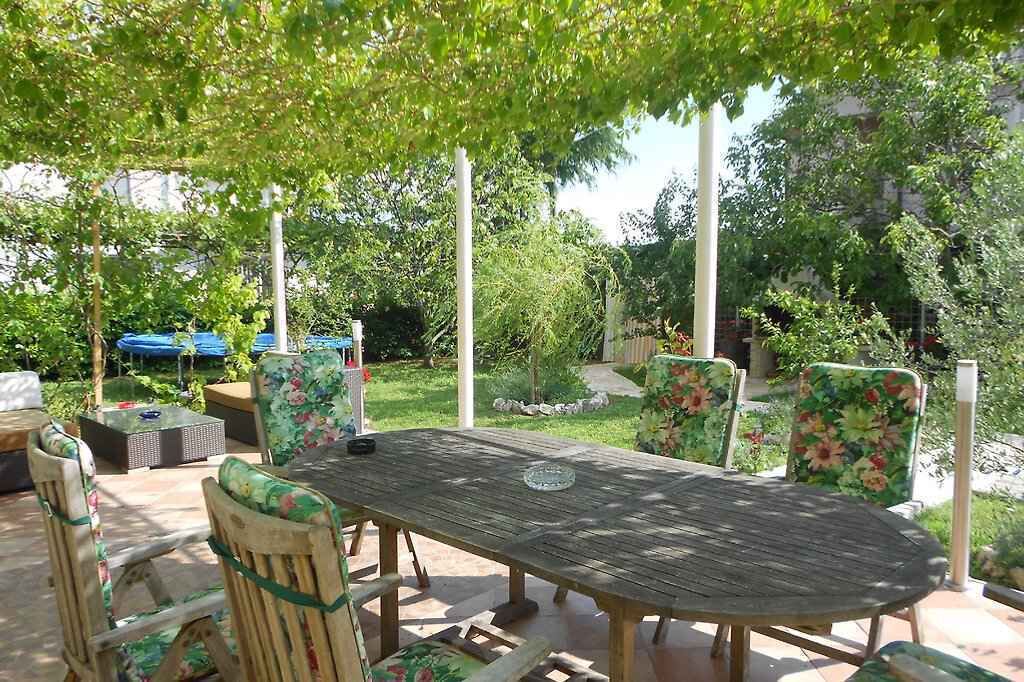 Une terrasse extérieure avec une table en bois et des chaises ornées de motifs floraux, entourée de verdure et bordée d’un chemin de pierres.