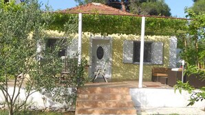 A yellow building with a red-tiled roof, a porch with a wooden chair, and a small garden area. - Holiday home 50 meters from the meter with large garden (Sukošan)